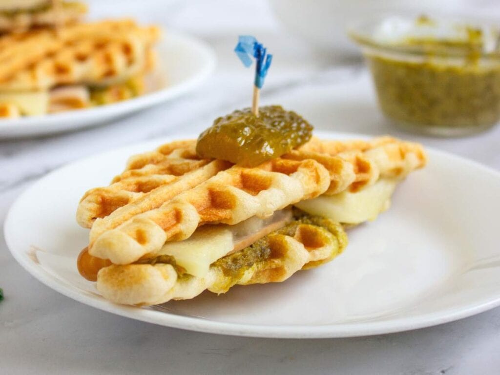 A waffle sandwich filled with melted cheese and sauce, topped with a pickle slice secured by a toothpick, served on a white plate.