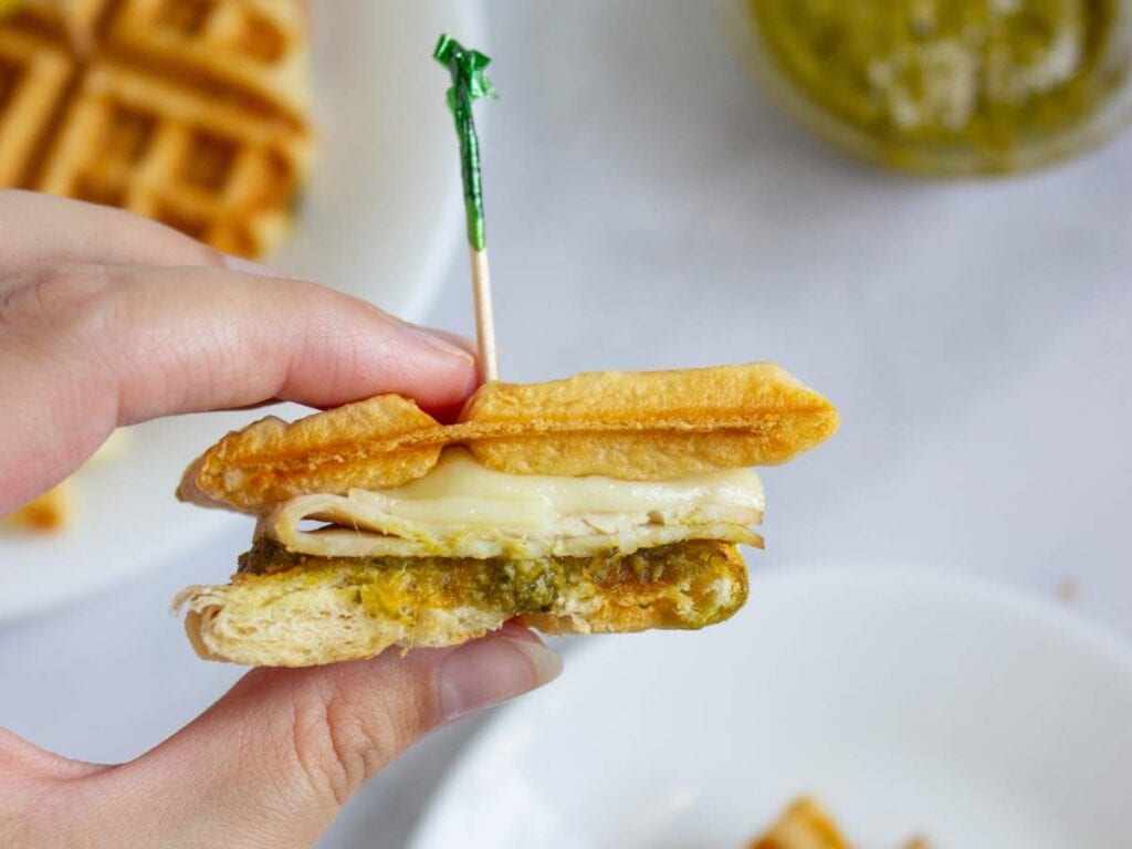 A hand holds a waffle sandwich with melted cheese and green pesto filling, secured by a toothpick.