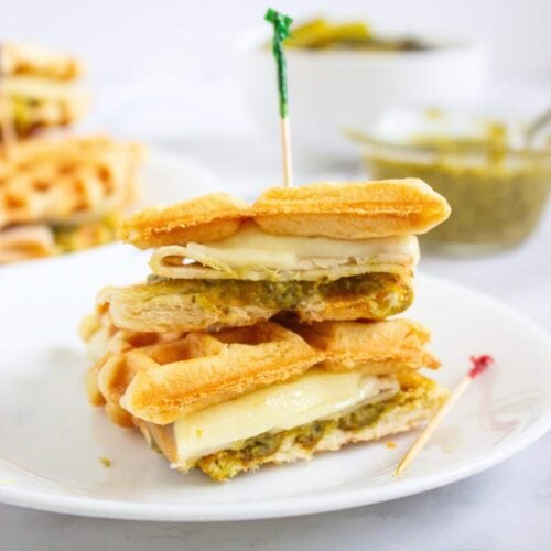 A sandwich made with waffles, melted cheese, and pesto is cut in half and stacked on a white plate with a toothpick holding it together. A bowl of pesto is in the background.