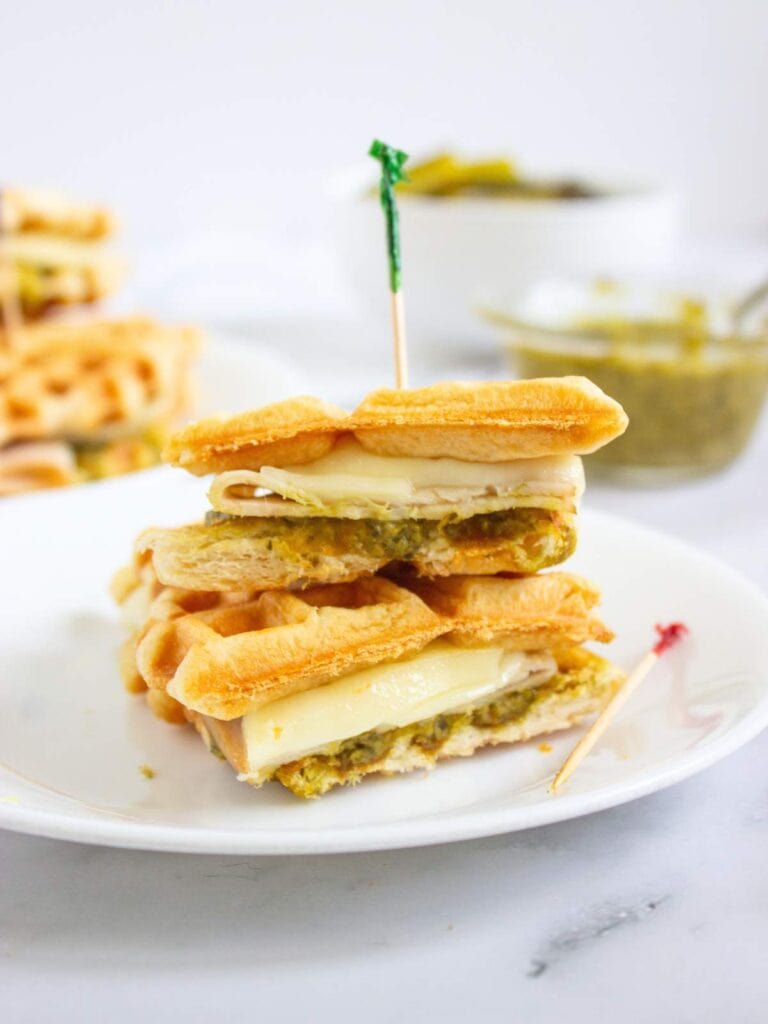 A sandwich made with waffles, melted cheese, and pesto is cut in half and stacked on a white plate with a toothpick holding it together. A bowl of pesto is in the background.