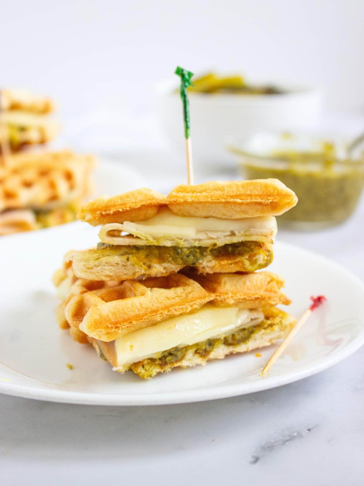 A sandwich made with waffles, melted cheese, and pesto is cut in half and stacked on a white plate with a toothpick holding it together. A bowl of pesto is in the background.