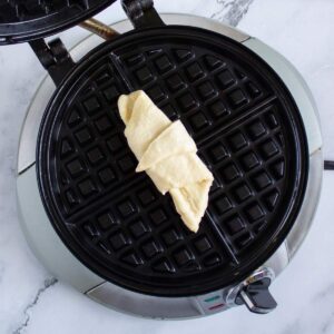 A raw crescent roll is placed on the center of an open, preheated waffle maker on a marble countertop.