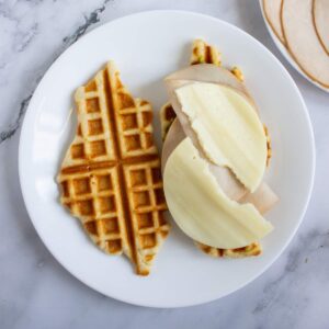 A plate with a broken waffle topped with slices of deli turkey and cheese, set on a marble surface.