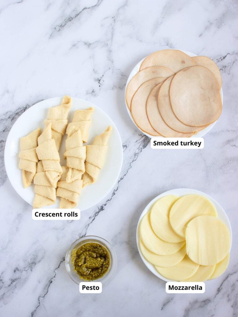 Three plates and a jar on a marble surface, containing crescent rolls, smoked turkey slices, mozzarella slices, and pesto. Each item is labeled.