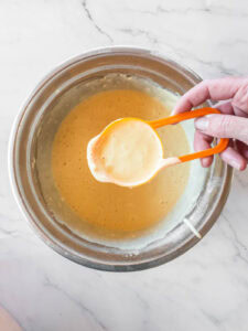 A hand holds a yellow measuring cup filled with pancake batter above a metal mixing bowl containing more batter on a marble countertop.