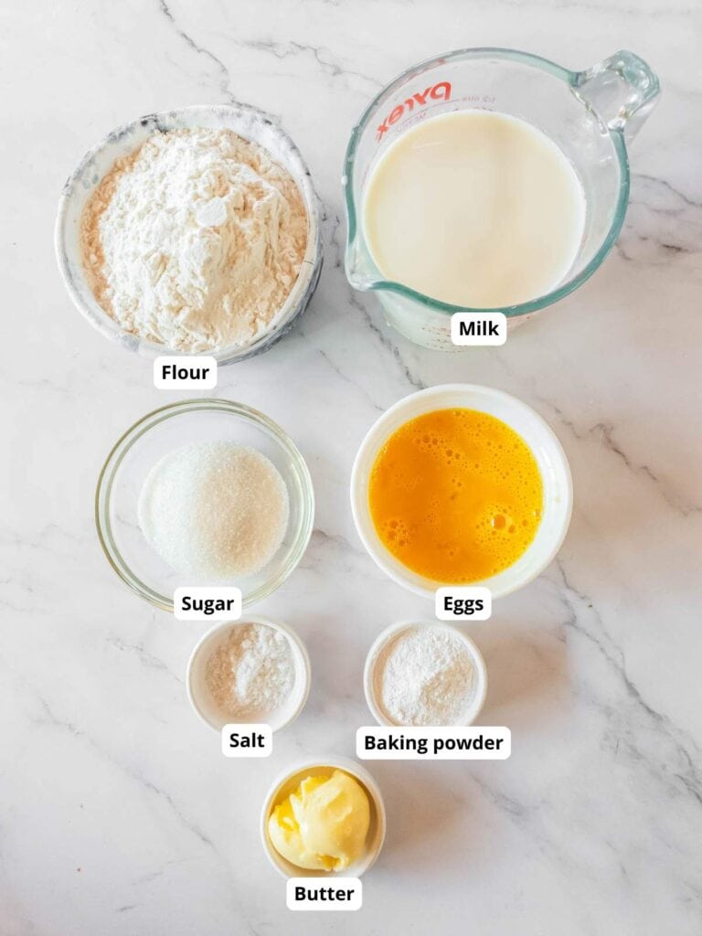 Top-down view of labeled baking ingredients on a marble surface: flour, milk, sugar, eggs, salt, baking powder, and butter.