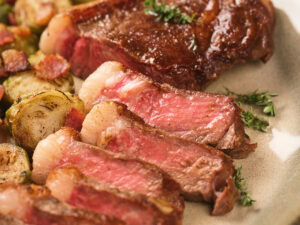 Sliced medium-rare steak on a plate, garnished with fresh herbs and served with roasted Brussels sprouts and bacon pieces.