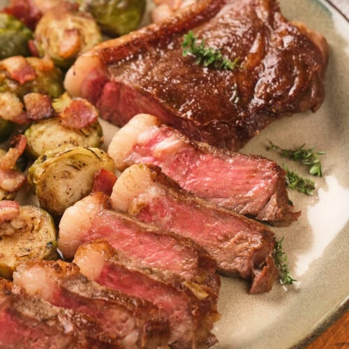Sliced medium-rare steak with seared crust served alongside roasted Brussels sprouts and bacon on a plate.