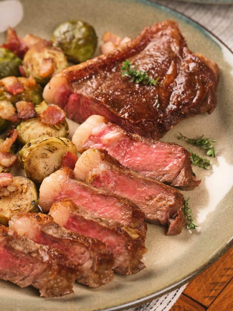 Sliced medium-rare steak with seared crust served alongside roasted Brussels sprouts and bacon on a plate.