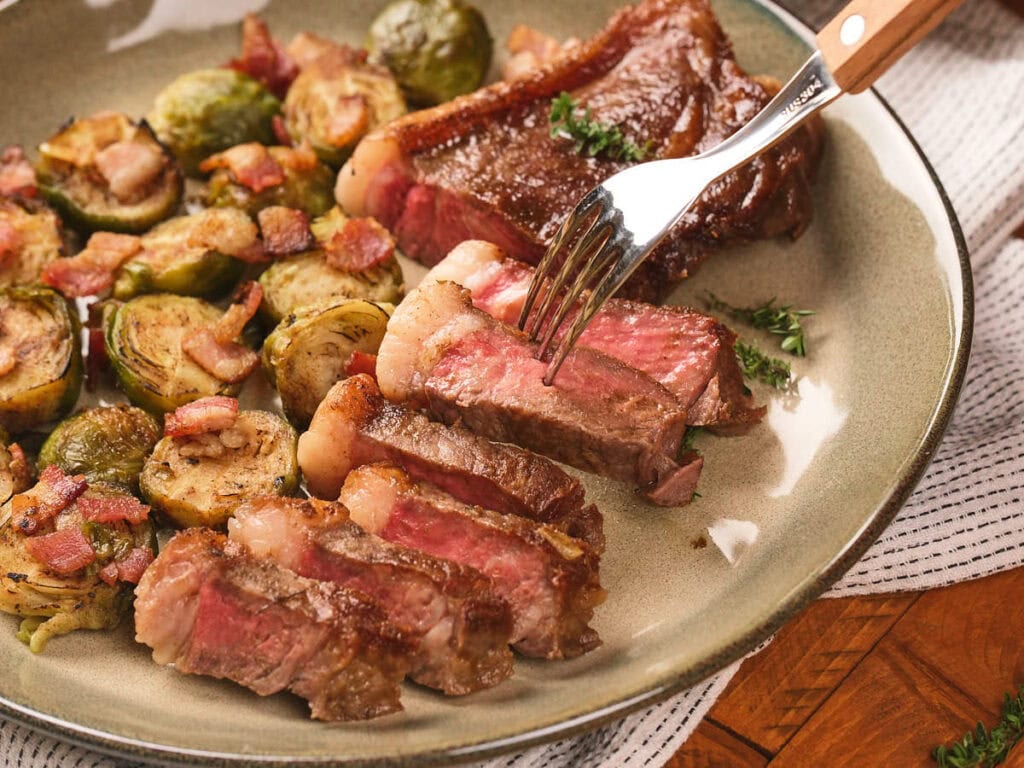 A plate with sliced medium-rare steak, roasted Brussels sprouts, and bacon pieces, with a fork holding a steak slice.