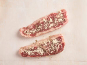 Two raw strip steaks are laid out on a light surface, each sprinkled with a mixture of seasoning and grated cheese.