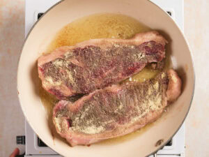 Two seasoned raw steaks are cooking in a beige skillet with melted butter or oil on a stovetop.