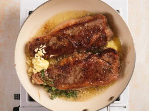 Two seared steaks cook in a pan with melted butter, chopped garlic, and fresh herbs on a stovetop.