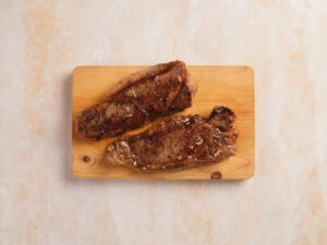 Two cooked steaks are placed side by side on a wooden cutting board against a light-colored background.