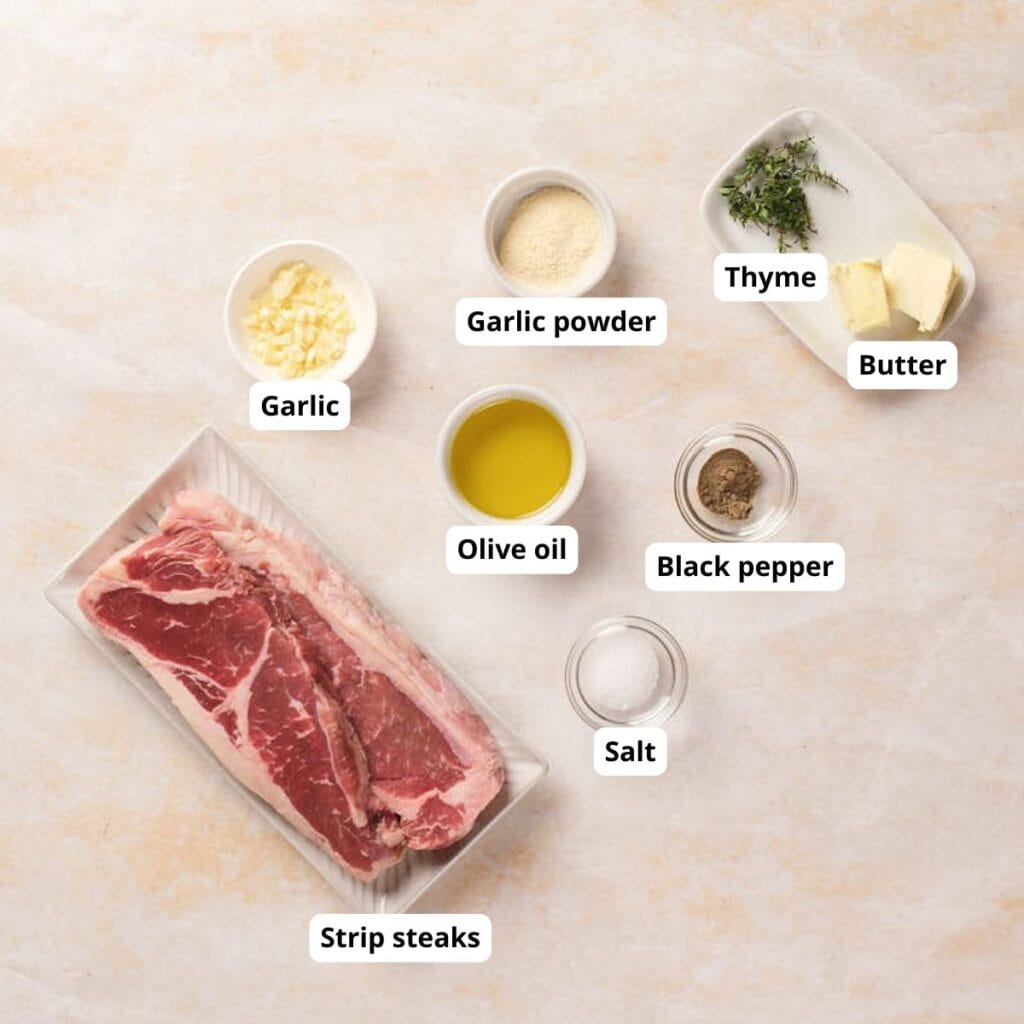 Labeled ingredients for cooking strip steaks, including garlic, garlic powder, thyme, butter, olive oil, black pepper, salt, and raw strip steaks on a light surface.