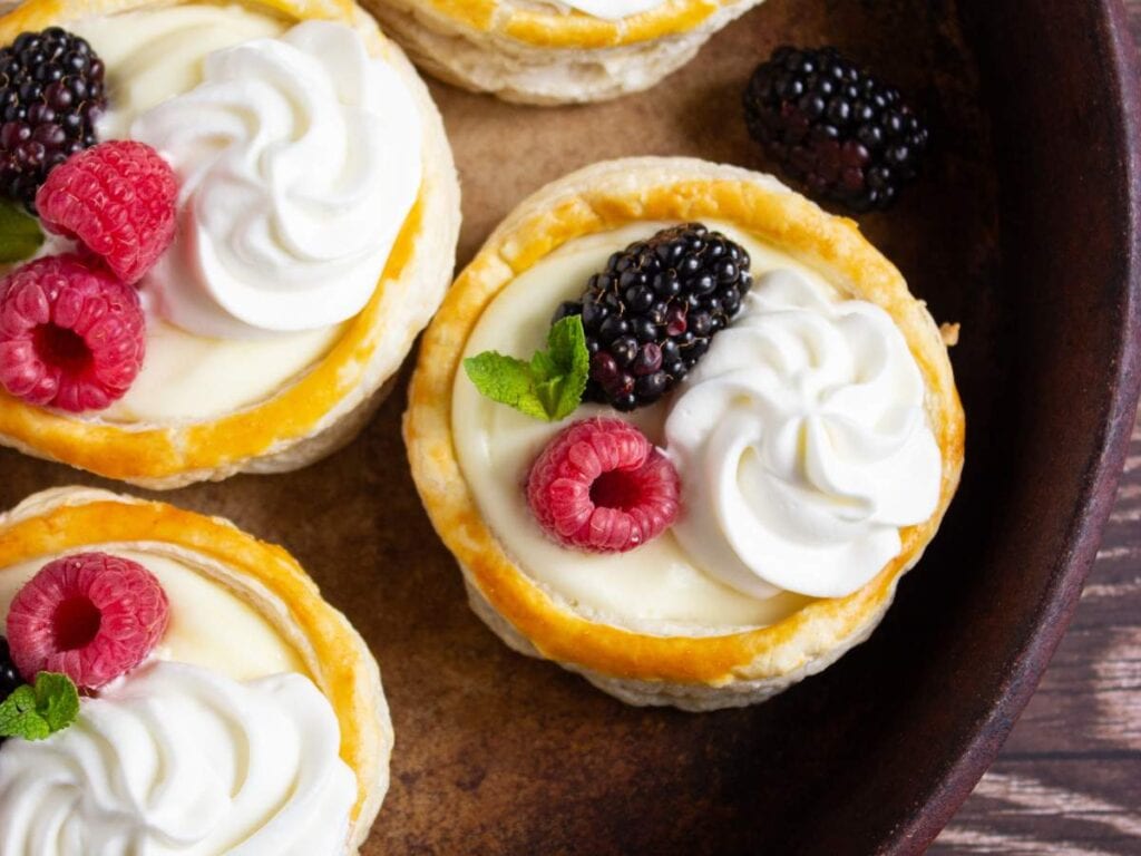 Pastry tarts filled with cream, topped with dollops of whipped cream, fresh raspberries, blackberries, and mint leaves, arranged on a brown plate.