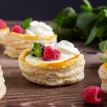 Round puff pastry shells filled with cream, topped with raspberries, blackberries, mint leaves, and a swirl of whipped cream, displayed on a wooden surface.