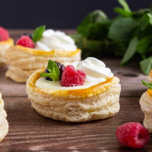 Round puff pastry shells filled with cream, topped with raspberries, blackberries, mint leaves, and a swirl of whipped cream, displayed on a wooden surface.