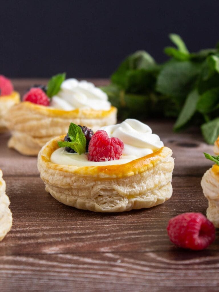 Round puff pastry shells filled with cream, topped with raspberries, blackberries, mint leaves, and a swirl of whipped cream, displayed on a wooden surface.