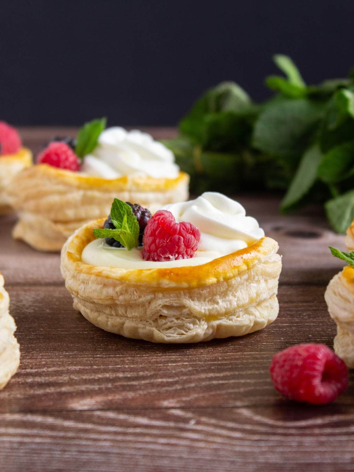 Round puff pastry shells filled with cream, topped with raspberries, blackberries, mint leaves, and a swirl of whipped cream, displayed on a wooden surface.