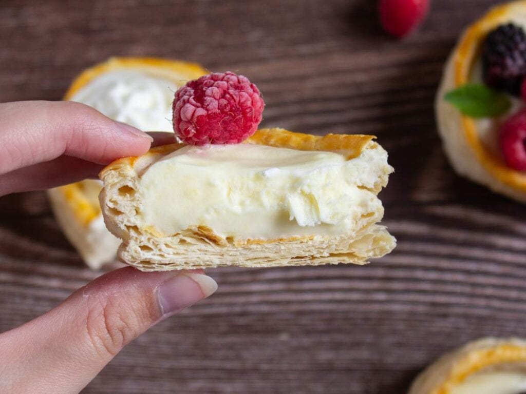 A hand holding a half-eaten pastry filled with cream and topped with a raspberry; other pastries with fruit are in the background on a wooden surface.