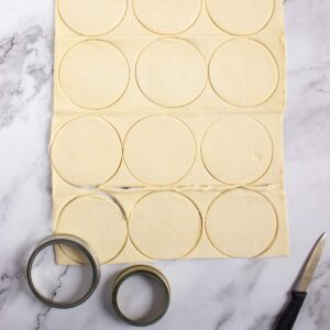 Sheet of dough with circular shapes cut out, two metal round cutters, and a paring knife on a marble surface.