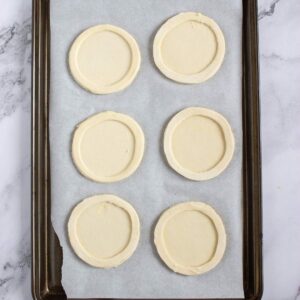 Six round, unbaked pastry circles with indented borders are arranged on a parchment-lined baking sheet.