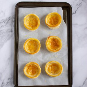 Six round, golden-brown pastry shells are arranged in two rows on a parchment-lined baking tray atop a marble surface.