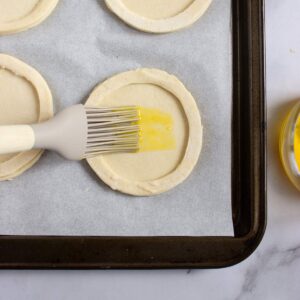 A pastry brush applies egg wash to round pieces of raw pastry dough on a baking tray lined with parchment paper.