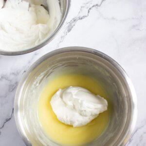 Two metal mixing bowls on a marble surface, one containing whipped cream and the other with a mixture of yellow batter and a dollop of whipped cream.