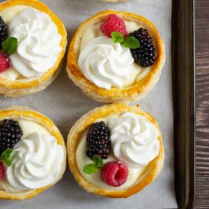 Four pastry tarts filled with cream, topped with whipped cream, blackberries, raspberries, and mint leaves on a baking tray lined with parchment paper.