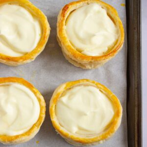 Four round puff pastry shells filled with creamy white custard are arranged on a parchment-lined baking sheet.