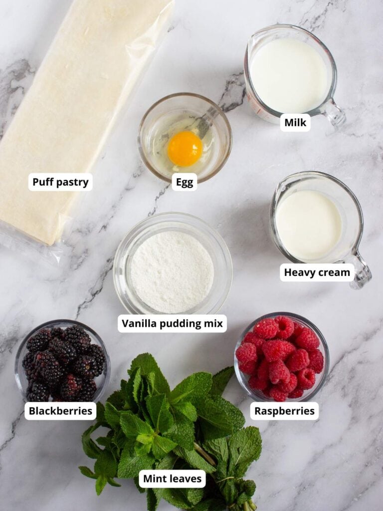 Ingredients for a dessert arranged on a marble surface: puff pastry, egg, milk, heavy cream, vanilla pudding mix, blackberries, raspberries, and mint leaves.