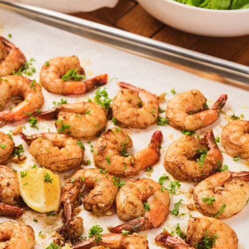 Seasoned shrimp garnished with chopped herbs and lemon wedges on a parchment-lined baking sheet, with a bowl of green salad in the background.