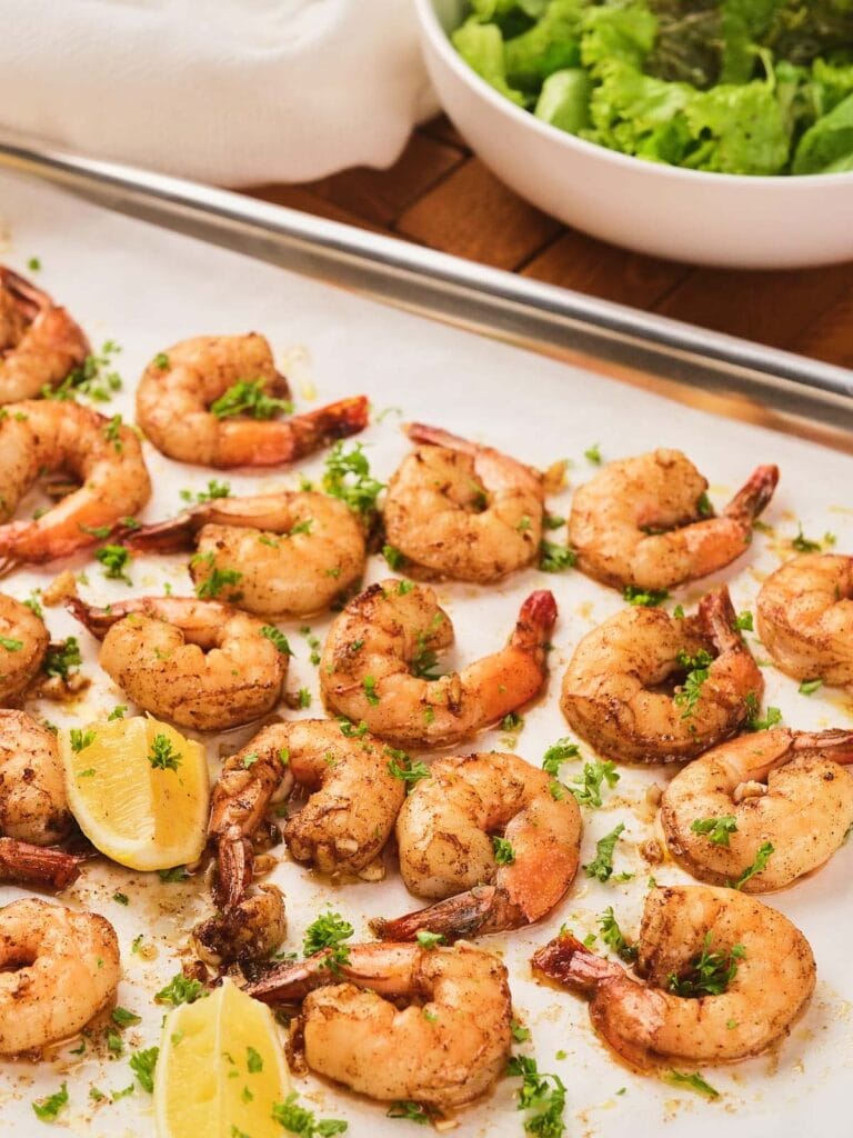 Seasoned shrimp garnished with chopped herbs and lemon wedges on a parchment-lined baking sheet, with a bowl of green salad in the background.