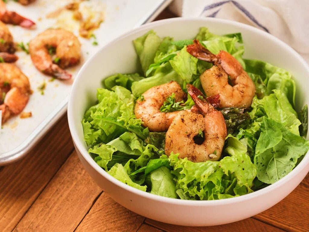 A white bowl filled with fresh green lettuce leaves and three seasoned, cooked shrimp.