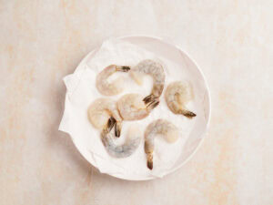 Six raw, peeled shrimp are arranged on a white paper towel-lined plate against a light background.
