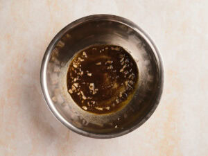 A metal mixing bowl containing a small amount of dark liquid with finely chopped garlic on a light-colored surface.