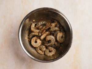 Raw shrimp marinated with spices and garlic in a stainless steel mixing bowl on a light-colored surface.