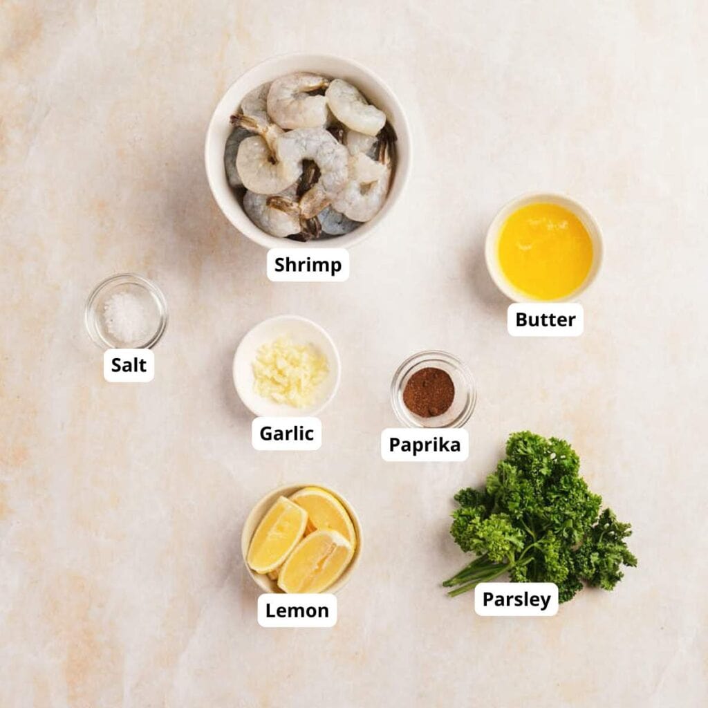 Top-down view of raw shrimp, melted butter, salt, minced garlic, paprika, lemon wedges, and parsley arranged on a light surface.