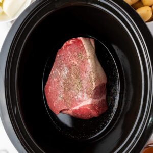 A raw seasoned beef roast sits in the center of a black slow cooker, with onions and potatoes visible nearby outside the cooker.