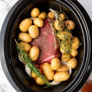 Uncooked pot roast with baby potatoes, carrots, onions, rosemary, and thyme arranged in a slow cooker.