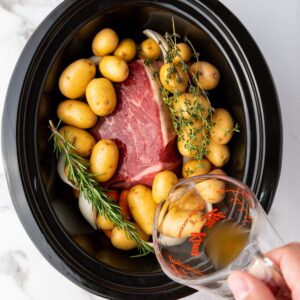 A raw beef roast, baby potatoes, rosemary, and thyme in a slow cooker with broth being poured from a measuring cup.