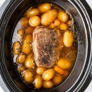 A slow cooker filled with a cooked beef roast, baby potatoes, carrots, broth, and sprigs of rosemary.