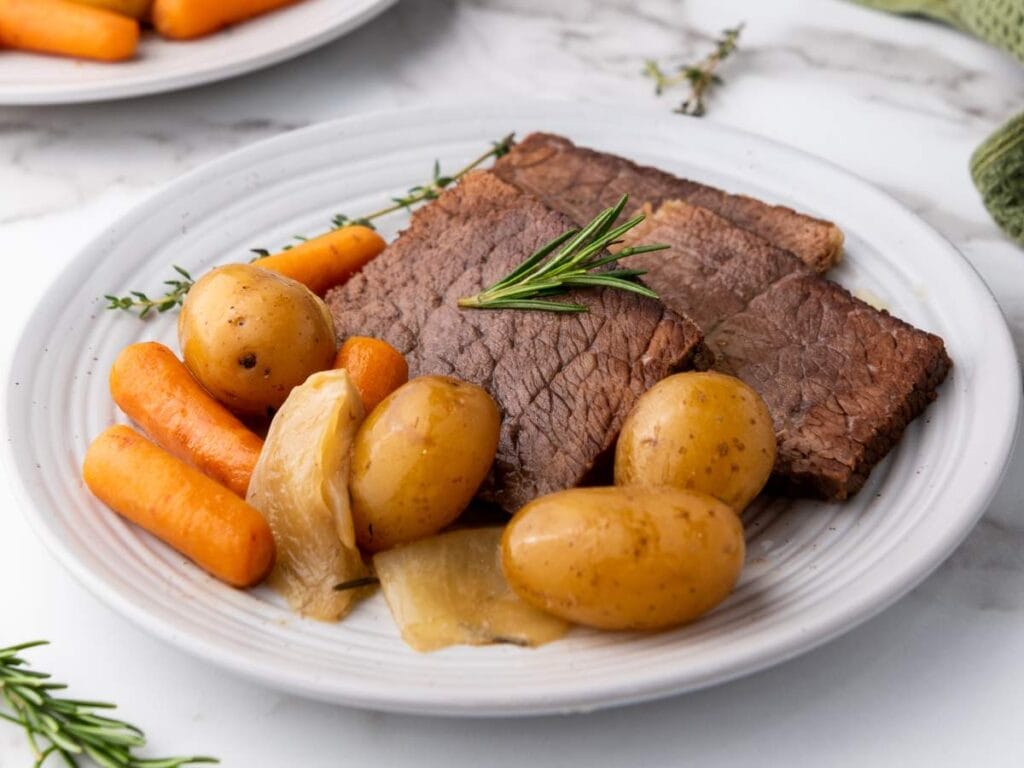 A plate with slices of roast beef, baby potatoes, carrots, and onion, garnished with a sprig of rosemary.