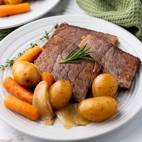 A plate with slices of roast beef, baby potatoes, carrots, and onions, garnished with a sprig of rosemary.