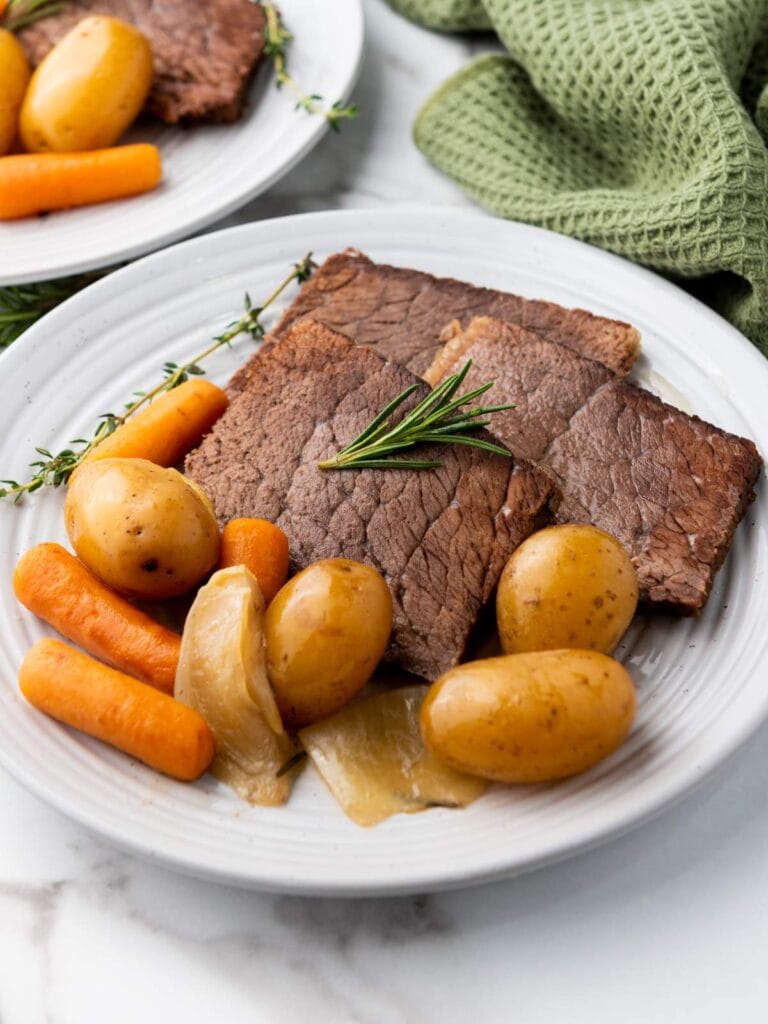 A plate with slices of roast beef, baby potatoes, carrots, and onions, garnished with a sprig of rosemary.
