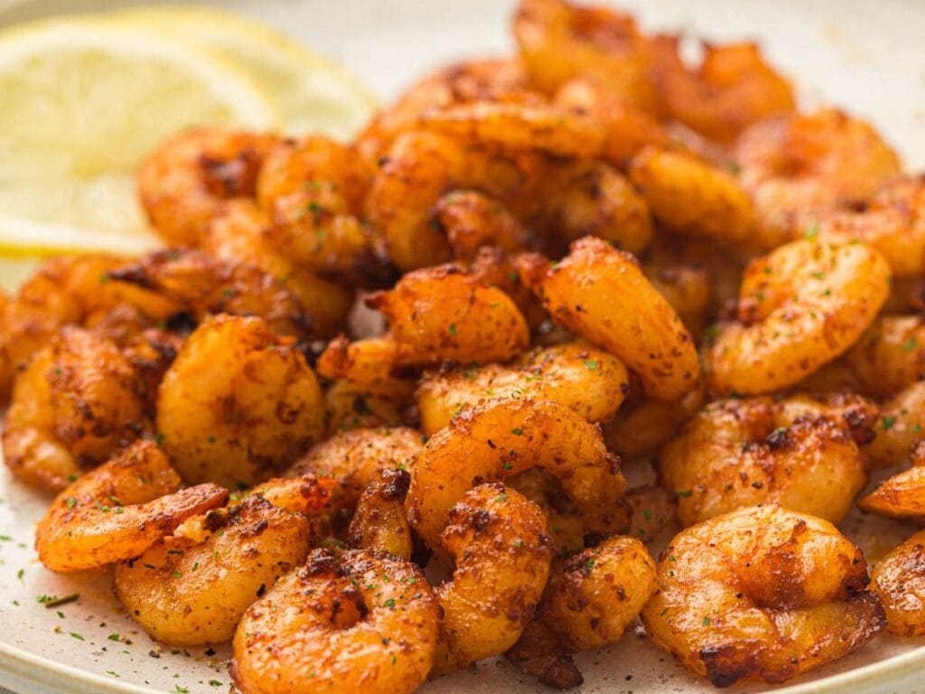 A plate of seasoned, cooked shrimp with two lemon slices in the background. The shrimp are golden brown and garnished with herbs.