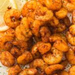 Seasoned cooked shrimp arranged on a plate with two slices of lemon in the corner, sprinkled with herbs.