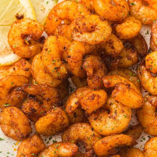 Seasoned cooked shrimp arranged on a plate with two slices of lemon in the corner, sprinkled with herbs.
